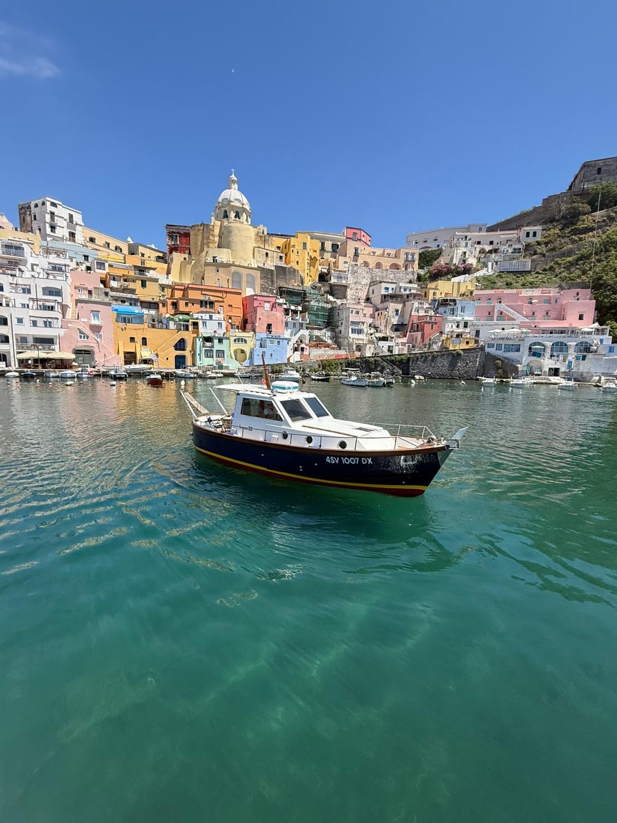 Wide view of the classic boat floating on emerald water beneath Procida's colourful waterfront
