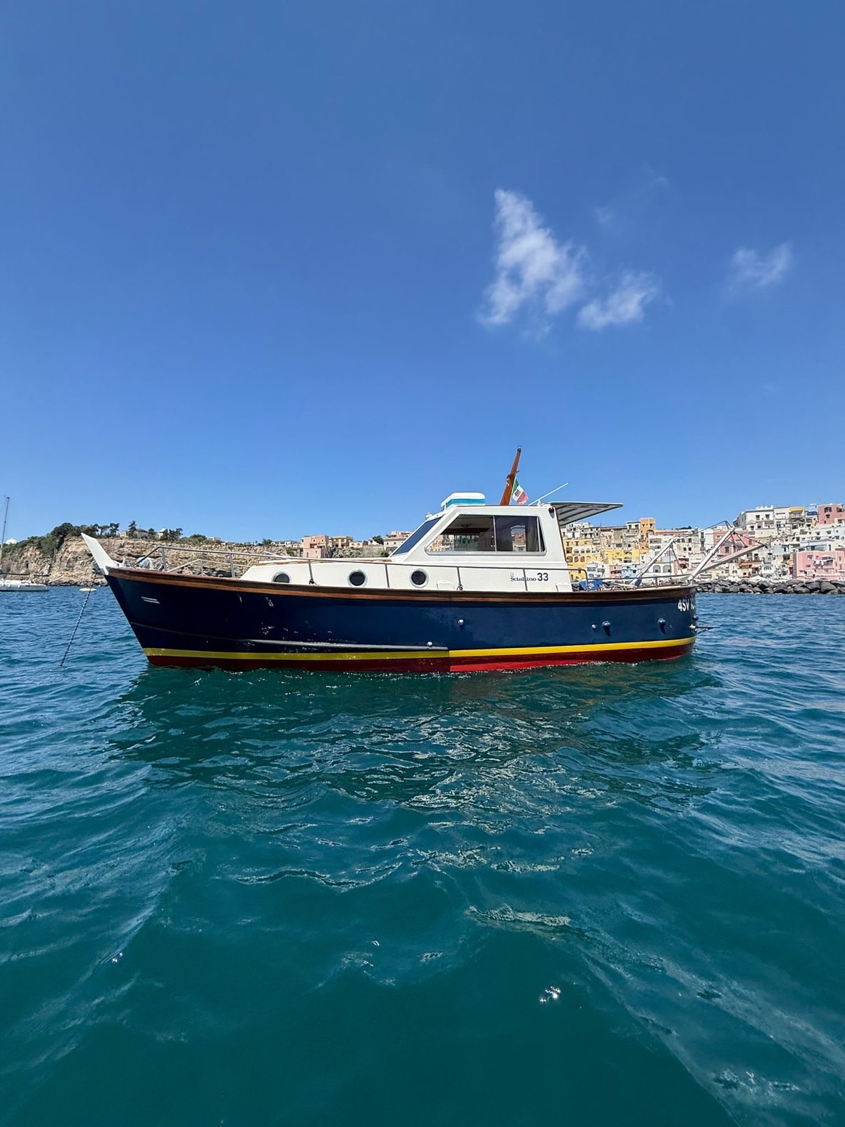Full profile of Il Lupo Cattivissimo II at anchor off Corricella, navy hull with yellow and red boot stripes