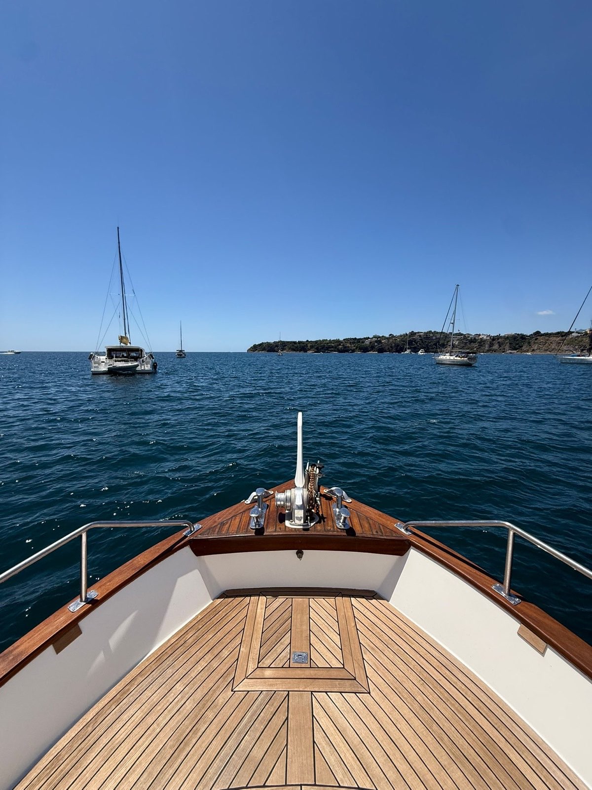 Teak foredeck with vintage windlass and cleats, open sea and sailboats of the Gulf of Naples ahead